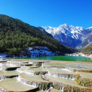 玉龍雪山白水台呈現層層堆疊的白色鈣化梯田景觀,清澈流水沿著階梯狀平台緩緩流下。遠方可見雪覆的玉龍雪山與翠綠山谷,藍天映襯壯麗自然景色,遊客在湖畔與平台附近欣賞玉龍雪山白水台獨特地貌。