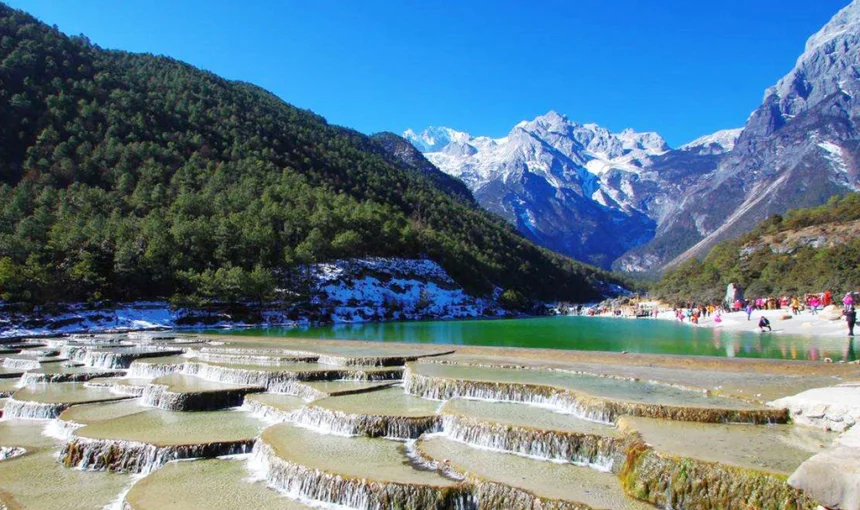 玉龍雪山白水台呈現層層堆疊的白色鈣化梯田景觀,清澈流水沿著階梯狀平台緩緩流下。遠方可見雪覆的玉龍雪山與翠綠山谷,藍天映襯壯麗自然景色,遊客在湖畔與平台附近欣賞玉龍雪山白水台獨特地貌。