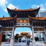 昆明金馬碧雞坊矗立於廣場,藍天下的傳統建築展現飛簷翹角與精美彩繪。牌坊刻有「金馬」字樣,與後方現代城市建築交織。