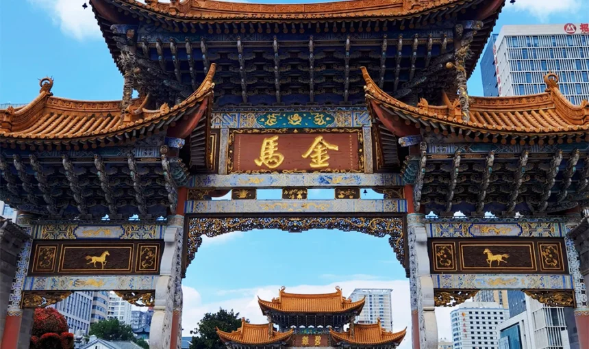 昆明金馬碧雞坊矗立於廣場,藍天下的傳統建築展現飛簷翹角與精美彩繪。牌坊刻有「金馬」字樣,與後方現代城市建築交織。