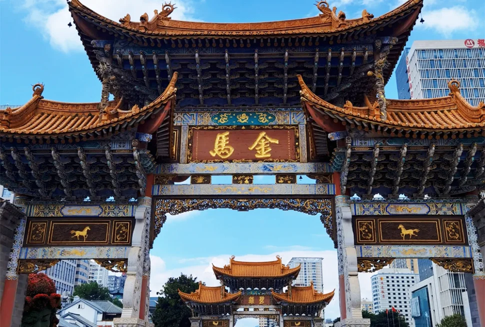 昆明金馬碧雞坊矗立於廣場，藍天下的傳統建築展現飛簷翹角與精美彩繪。牌坊刻有「金馬」字樣，與後方現代城市建築交織。