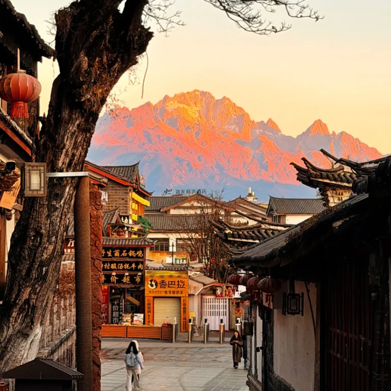清晨的 麗江古城,傳統瓦屋、靜謐石板巷道,遠方是被晨光染金的玉龍雪山。