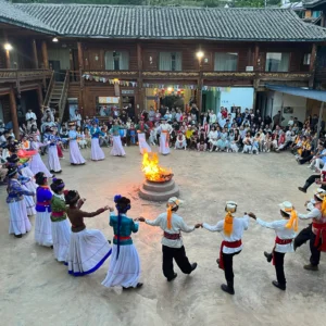 瀘沽湖篝火晚會。身穿傳統摩梭服飾的男女手拉手圍成一圈，繞著巨大的點燃火坑起舞，身後是落座的觀眾和一座多層木建築。