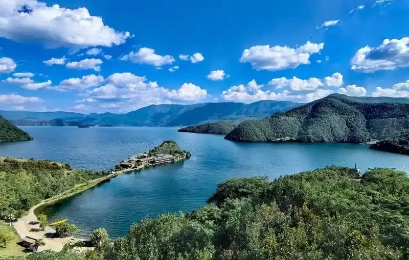 瀘沽湖壯麗湖景在晴朗藍天下展開,湖水深藍清澈,群山環繞湖泊。湖中央的小島與連接岸邊的堤道清晰可見,周圍綠意盎然的森林與遠方山巒層層延伸,白雲點綴天空,展現瀘沽湖高原湖泊寧靜而壯觀的自然風光。