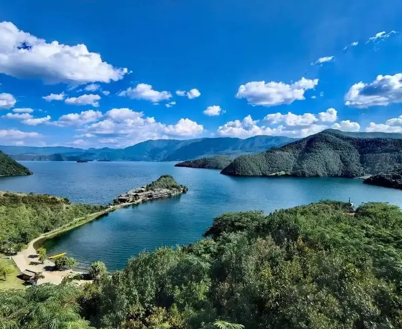 瀘沽湖壯麗湖景在晴朗藍天下展開，湖水深藍清澈，群山環繞湖泊。湖中央的小島與連接岸邊的堤道清晰可見，周圍綠意盎然的森林與遠方山巒層層延伸，白雲點綴天空，展現瀘沽湖高原湖泊寧靜而壯觀的自然風光。