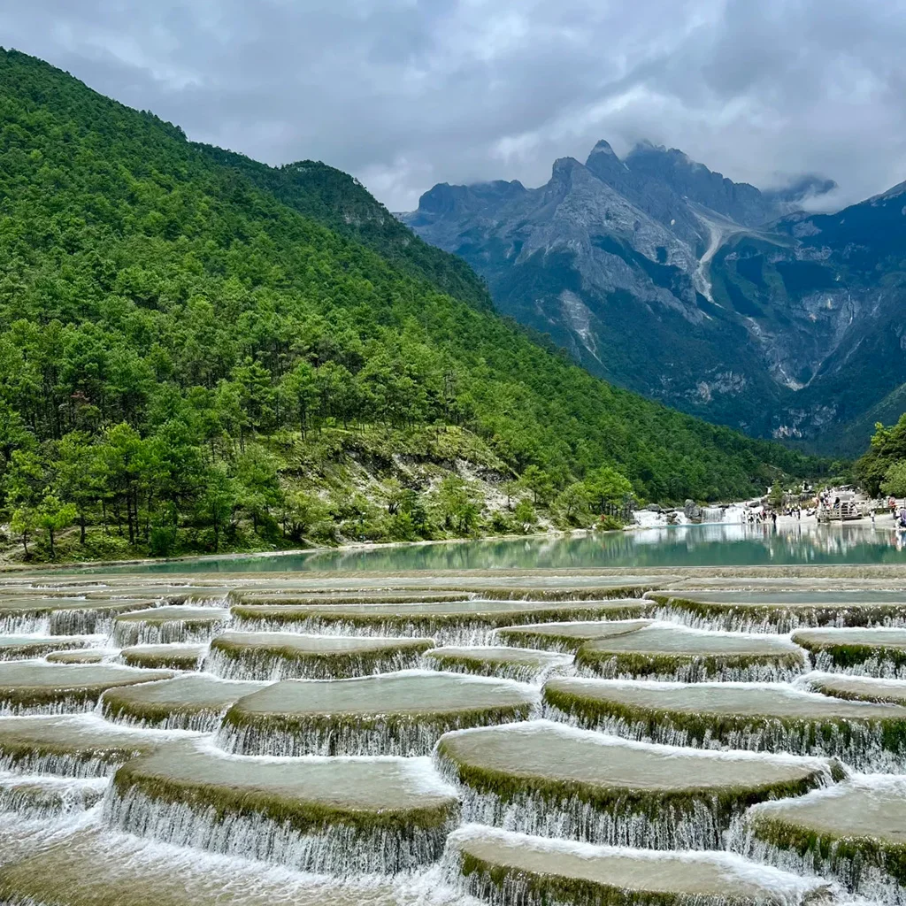 雲南麗江玉龍雪山下的藍月谷(白水河)絕美景致,層層疊疊的碧綠瀑布沿著石灰岩台階傾瀉,背景是雪山與翠綠森林。