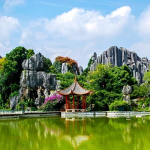 昆明石林景區內的奇特石灰岩地貌聳立於綠樹之間,湖面前方有一座中式涼亭倒映在水中。鋸齒狀石峰與翠綠植被相互交織,在藍天白雲映襯下展現昆明石林獨特壯觀的喀斯特地貌與自然景觀。