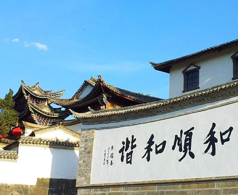 雲南騰衝的 和順古鎮 古建築景觀。白色石牆上寫著「和順和諧」書法字樣，牆後可見傳統中式屋頂與飛簷建築，青瓦層疊，展現典型的古鎮風格。晴朗藍天下，古鎮街巷寧靜雅致，整體畫面呈現濃厚的歷史文化氣息與雲南傳統人文風貌。