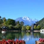 雲南麗江的 黑龍潭 湖景與遠方的 玉龍雪山 同框入鏡。清澈湖水倒映著雪山、藍天與周圍的綠樹景觀,湖畔可見傳統中式亭子與古建築,景色寧靜而壯麗。前景鮮豔花卉點綴湖岸,使整個畫面呈現出層次豐富且富有詩意的自然與人文景觀。
