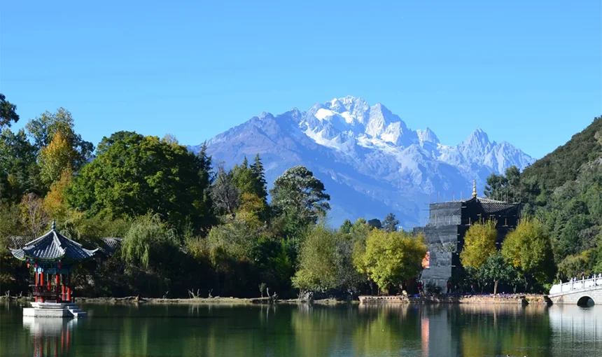 雲南麗江的 黑龍潭 湖景與遠方的 玉龍雪山 同框入鏡。清澈湖水倒映著雪山、藍天與周圍的綠樹景觀,湖畔可見傳統中式亭子與古建築,景色寧靜而壯麗。前景鮮豔花卉點綴湖岸,使整個畫面呈現出層次豐富且富有詩意的自然與人文景觀。