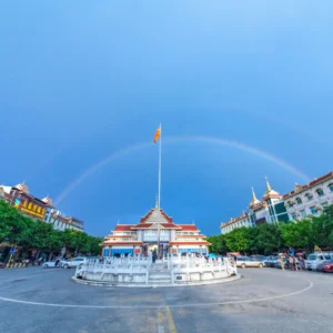姐告口岸廣場景觀，中央設有圓形噴泉與高聳旗桿，周圍為具有東南亞風格的建築群與商店街。天空出現一道完整彩虹橫跨廣場上方，與藍天形成壯麗畫面，展現雲南邊境城市熱鬧而獨特的口岸景觀與旅遊氛圍。