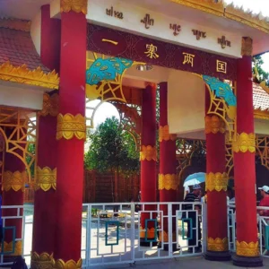 一寨兩國景區入口建築，紅色立柱與金色裝飾呈現東南亞民族風格，上方牌匾寫著「一寨兩國」，遊客在入口處參觀拍照，展現中緬邊境一寨兩國獨特的地理與文化景觀。