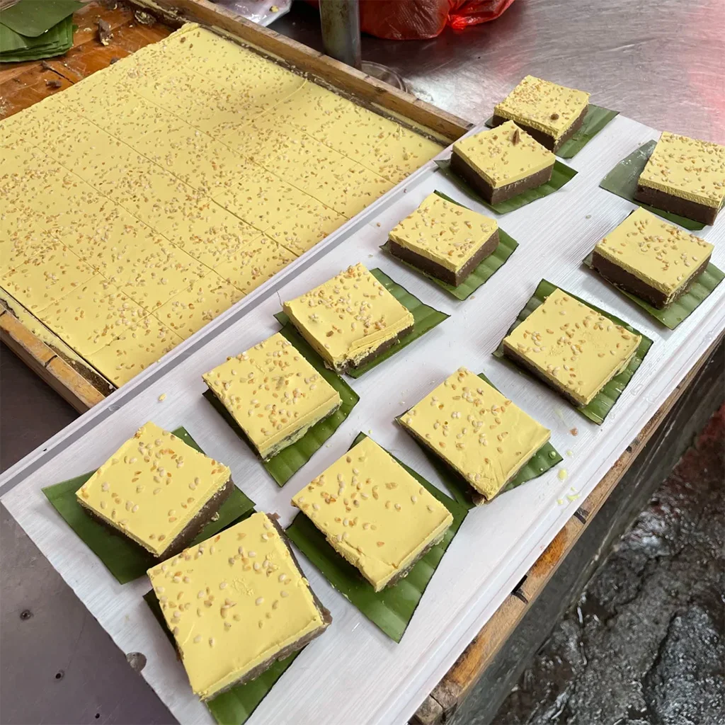 地道騰衝桂花蛋糕,雲南特色甜點。金黃色方形糕點灑滿芝麻,內餡夾層為紅豆沙,置於芭蕉葉上,展現傳統在地手藝