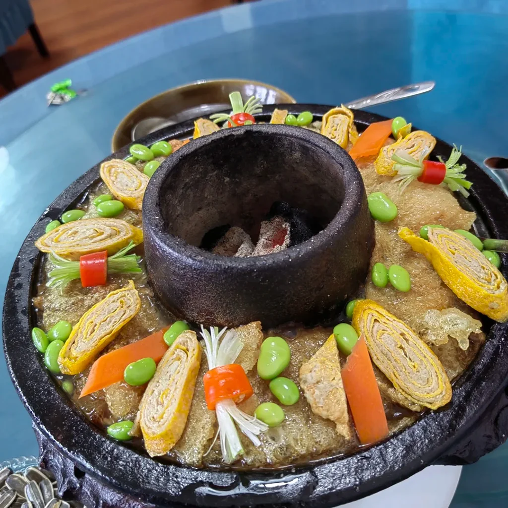 地道騰衝土鍋子,雲南傳統火鍋特色料理。手工黑陶鍋內鋪滿蛋卷、毛豆、炸豬皮,以濃郁湯頭慢燉。