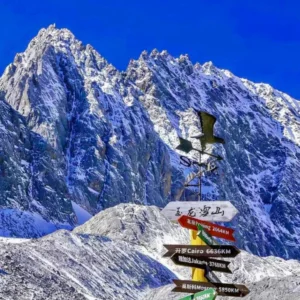 雲南麗江著名的 玉龍雪山 壯麗雪峰景觀。高聳陡峭的雪山覆蓋著白雪,在湛藍天空下顯得格外壯觀。前景是一個寫著「玉龍雪山」的方向指示牌與風向標,指向世界多個城市距離,為畫面增添旅遊地標特色,展現出這座著名雪山的雄偉與魅力。