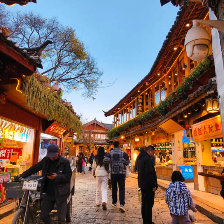 雲南著名歷史景區 麗江古城 的傍晚街景。石板路兩側是傳統木質建築與商店,屋簷掛著燈飾與招牌。遊客與當地居民在古街上漫步購物,街道燈光溫暖,營造出熱鬧又富有文化氣息的古鎮氛圍,展現麗江古城獨特的旅遊魅力