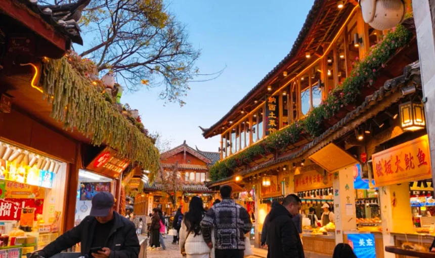 雲南著名歷史景區 麗江古城 的傍晚街景。石板路兩側是傳統木質建築與商店,屋簷掛著燈飾與招牌。遊客與當地居民在古街上漫步購物,街道燈光溫暖,營造出熱鬧又富有文化氣息的古鎮氛圍,展現麗江古城獨特的旅遊魅力