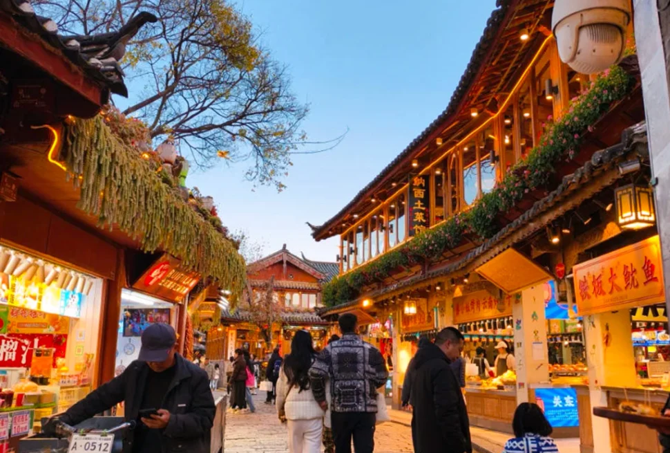 雲南著名歷史景區 麗江古城 的傍晚街景。石板路兩側是傳統木質建築與商店，屋簷掛著燈飾與招牌。遊客與當地居民在古街上漫步購物，街道燈光溫暖，營造出熱鬧又富有文化氣息的古鎮氛圍，展現麗江古城獨特的旅遊魅力