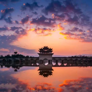 建水雙龍橋夕陽景觀，古老石拱橋橫跨水面，橋中央的傳統亭閣在晚霞映照下形成優美剪影，橋洞與天空雲彩倒映在平靜水面上，呈現雙龍橋典雅壯麗且充滿歷史韻味的古橋風光
