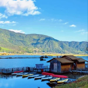 騰沖北海濕地自然景觀，湖面平靜映照藍天與群山，岸邊木屋與棧道延伸至水面，小船整齊停泊於碼頭旁。遠方青山環繞濕地湖泊，展現騰沖北海濕地獨特的高原湖泊生態與寧靜優美的自然風光
