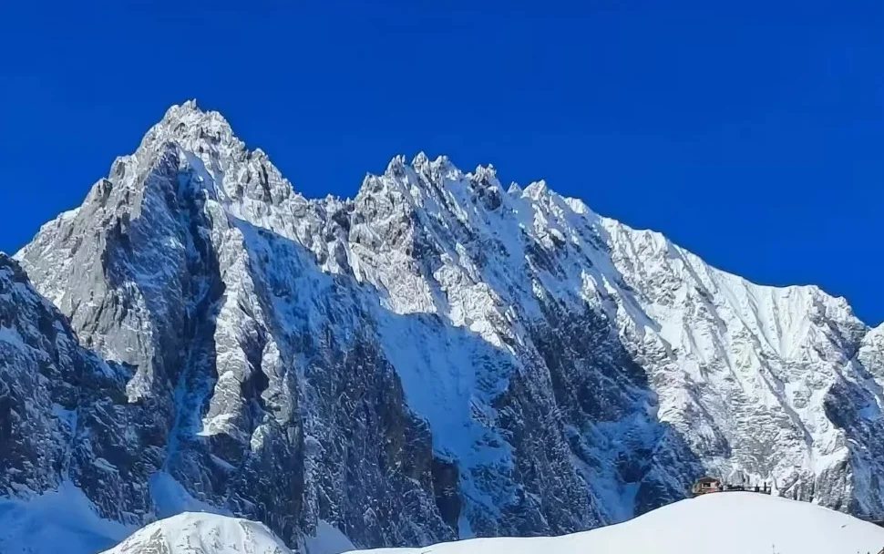 雲南麗江玉龍雪山，雄偉鋸齒狀積雪山峰聳立於澄澈深藍天空下，山腳有小型觀景平台。