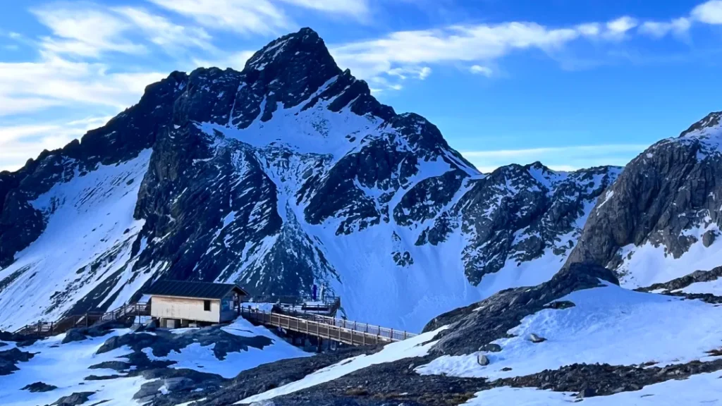 雲南麗江玉龍雪山,雄偉積雪山峰拔地而起,山腳有木棧道與山中小屋,背景是澄澈藍天。