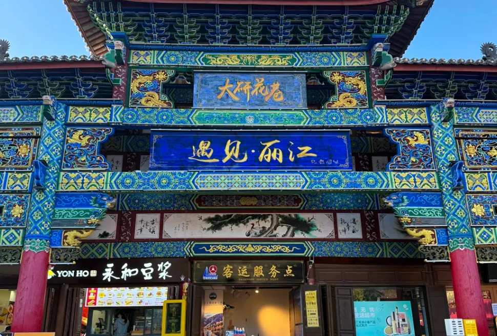 雲南麗江古城大研花巷，雕梁畫棟的傳統中式牌樓，「遇見・麗江」字樣，下方是在地店家，背景是澄澈藍天。