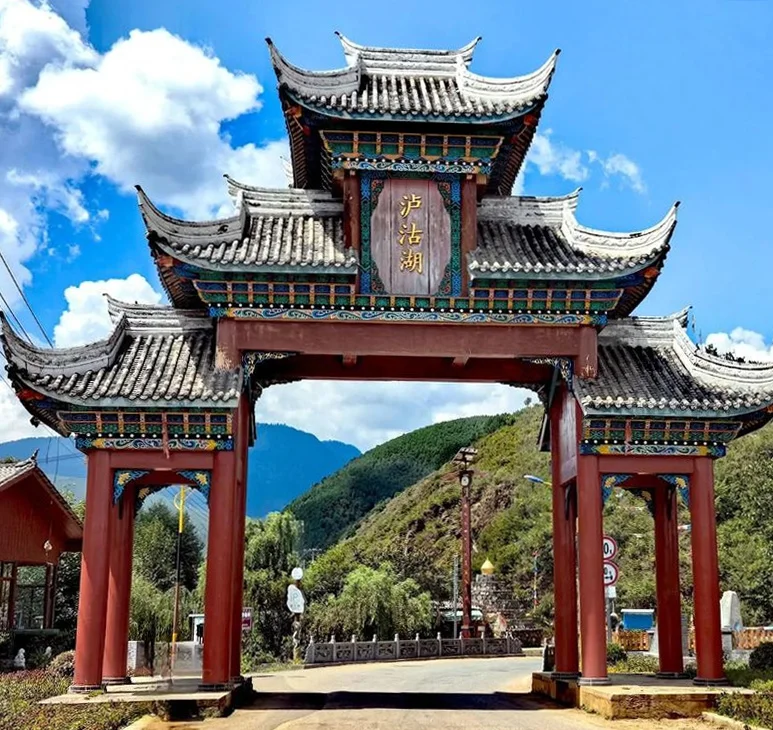 瀘沽湖景區入口的傳統中式牌樓矗立在道路上方,紅色立柱與多層飛簷屋頂展現典雅建築風格。牌樓中央寫著「瀘沽湖」三字,背後是綠色山巒與藍天白雲,呈現通往瀘沽湖自然風景區的地標入口與濃厚地方特色。