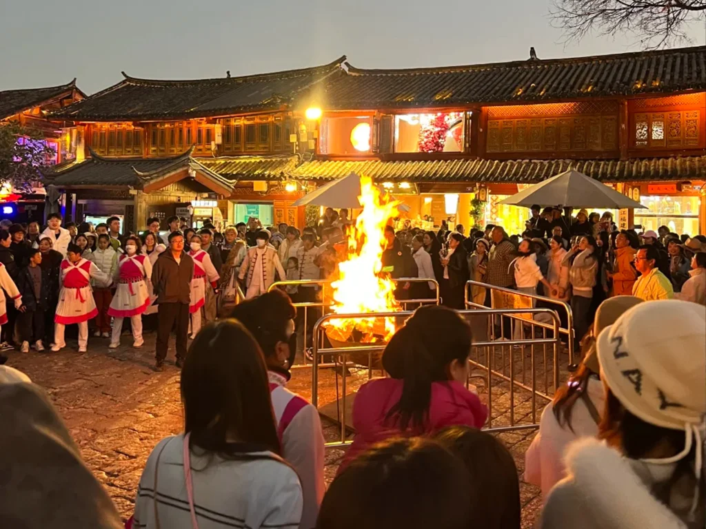 雲南麗江古城四方街篝火晚會,群眾圍繞熊熊篝火,背景是傳統納西建築與暮色天空。