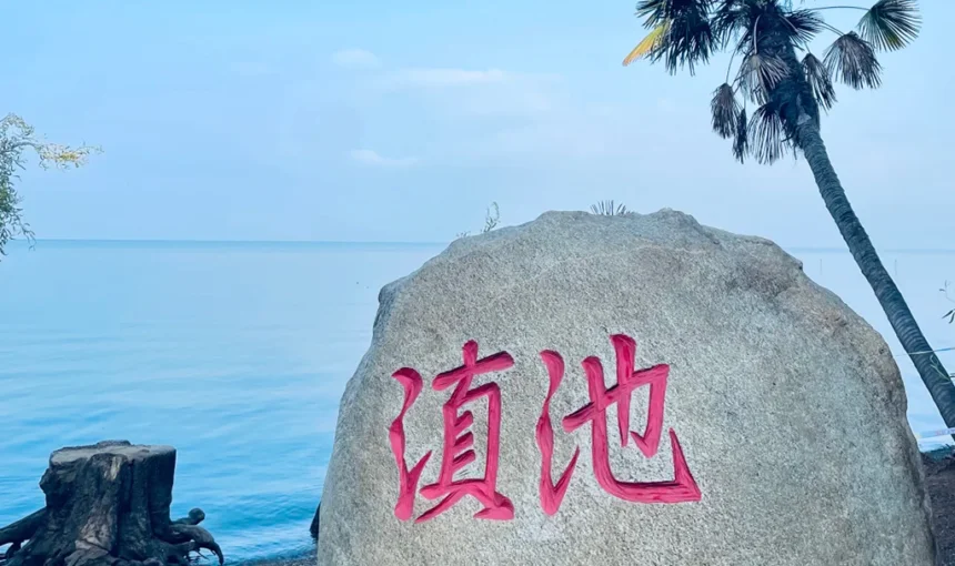 昆明滇池的經典地標石碑,刻有紅色「滇池」大字的灰色石頭,背景是湛藍的湖水與藍天,旁邊還有棕櫚樹點綴。