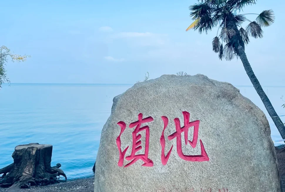 昆明滇池的經典地標石碑，刻有紅色「滇池」大字的灰色石頭，背景是湛藍的湖水與藍天，旁邊還有棕櫚樹點綴。