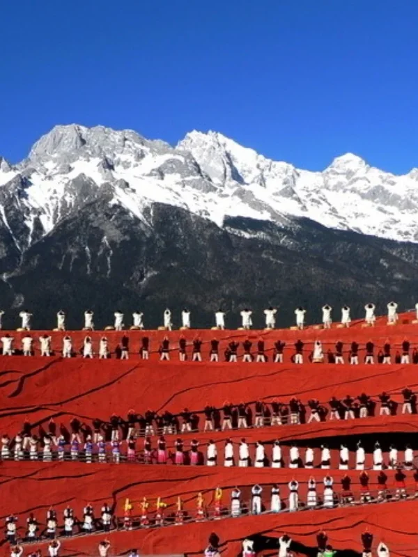 印象麗江大型實景表演在玉龍雪山下上演,紅色階梯舞台上眾多演員身著民族服飾整齊排列演出,背後是壯麗的雪山與藍天景觀,展現麗江獨特的民族文化與印象麗江震撼的戶外表演場面