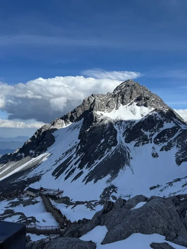 雲南麗江玉龍雪山,雄偉雪山聳立於澄澈藍天之下,木棧道沿著岩坡蜿蜒向上,盡顯高原壯麗風光。