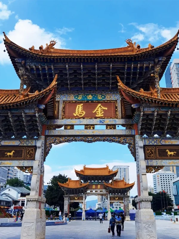 昆明金馬碧雞坊矗立於廣場,藍天下的傳統建築展現飛簷翹角與精美彩繪。牌坊刻有「金馬」字樣,與後方現代城市建築交織。