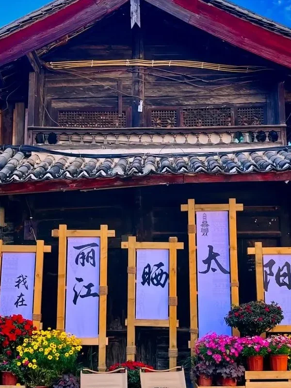麗江古城傳統木造建築外觀,屋頂覆蓋灰色瓦片,展現納西族古城建築特色。門前擺放花卉盆栽與木製展示架,上方書寫「我在麗江晒太陽」字樣,呈現悠閒的古城生活氛圍,是遊客拍照與體驗麗江古城文化的熱門場景。