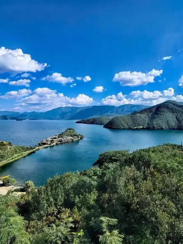瀘沽湖壯麗湖景在晴朗藍天下展開,湖水深藍清澈,群山環繞湖泊。湖中央的小島與連接岸邊的堤道清晰可見,周圍綠意盎然的森林與遠方山巒層層延伸,白雲點綴天空,展現瀘沽湖高原湖泊寧靜而壯觀的自然風光。
