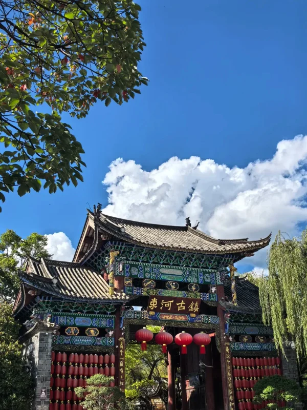 麗江束河古鎮的牌樓,彩繪木雕與紅燈籠相映,藍天白雲下,是古鎮最具儀式感的入口畫面。