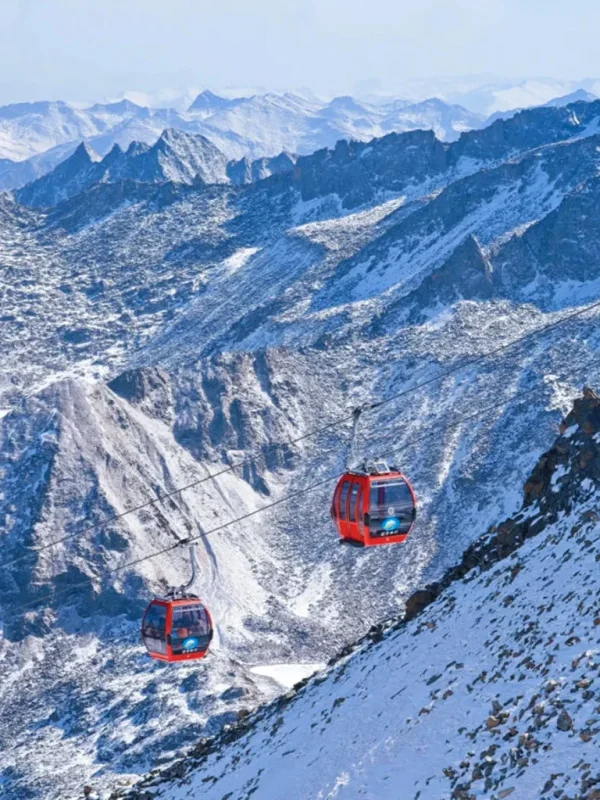 雲南麗江玉龍雪山冰川公園大索道的高山纜車景觀。紅色纜車在高空運行,穿越壯麗的雪山與冰川山脈,遠處連綿山峰覆蓋著白雪。崎嶇山脊與冰雪地貌展現高原壯觀景色,整個畫面呈現出震撼的自然風光與著名的高山旅遊體驗。